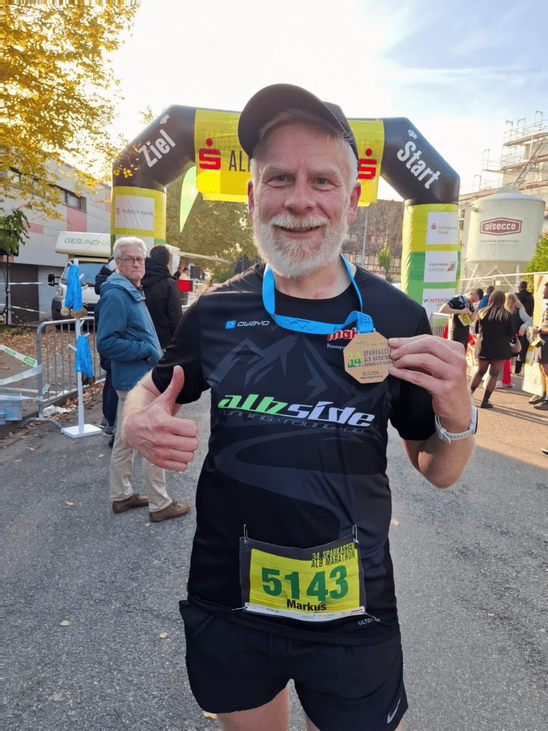 Marathon runner with bib number 5143, smiling and giving a thumbs-up at a race finish line.