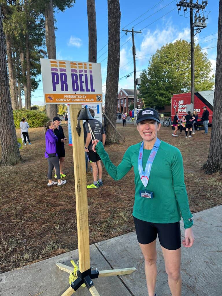 Elke woman in running gear with medal, standing next to race bell at event.